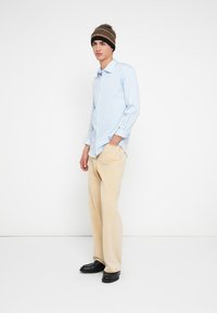 Svetlo modra srajca z gumbi s črtami, bež korduro hlače in temno rjava pletena kapa s črtami. Črni čevlji. Položaj stoje.