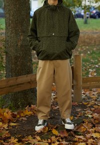 Personne debout en plein air sur des feuilles tombées, portant une veste verte à motifs, un pantalon beige et des baskets vertes et blanches, les mains dans les poches.