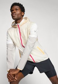 Giacca leggera in crema con cappuccio, zip rose e dettagli gialli. Presenta una tasca sul petto ed è abbinata a pantaloni sportivi neri.