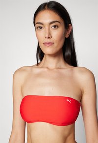Červený bandeau bikini top vyrobený z hladkej látky, s naberanými bokmi a malým bielym logom vpredu. Bez ramienok.