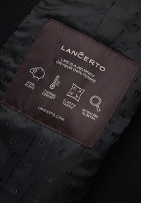 Czarna tkanina z subtelnym wzorem posiada brązową metkę. Metka zawiera ikony oznaczające mieszankę wełny, komfort termiczny, elastyczny materiał oraz dbałość o szczegóły.