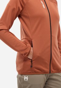 Veste à coque souple en orange brûlé avec un tissu texturé, dotée d'une fermeture éclair à l'avant, de deux poches latérales et d'un logo au bas.