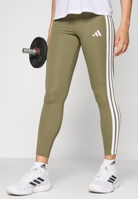 Leggings sportivi verde oliva con strisce laterali bianche, vestibilità slim e una texture liscia, abbinati a sneaker bianche. Un manubrio nero è tenuto in una mano.