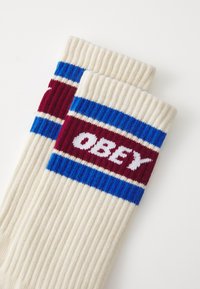 Ponožky po kolená v krémovej farbe s ryhovanou textúrou. Majú červené, modré a biele pruhy a logo "OBEY" v bielej farbe na bordovom pruhu.