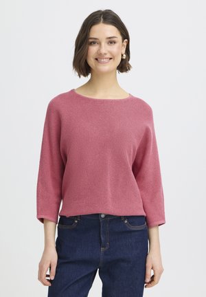 Lühikeste tumedate juustega naine, kes kannab roosat pika varrukaga kampsunit ja tumeid siniseid teksapükse, naeratades ühevärvilise valge tausta ees.