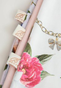 Gėlėmis raštuotas audinių gabalėlių krūva su rožinėmis kraštinėmis ir auksu siuvinėtais ženkliukais „BAKER“ ant šviesios fono.