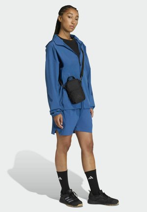 Niebieski komplet sportowy: lekka kurtka i spodenki. Do tego czarne buty i skarpetki z białymi logo. Mała czarna torba na ramię.