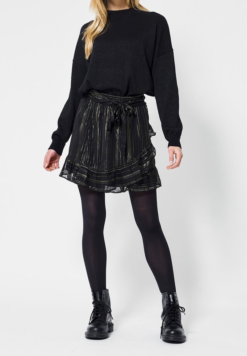 Černý pletený svetr s volným střihem, spárovaný s černou sukní s zlatými pruhy a volánkovým lemem. Noseno s černými punčochami a šněrovacími botami.