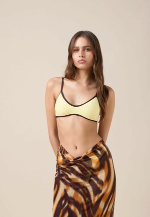 Młoda kobieta o długich brązowych włosach, ubrana w żółty top bikini z czarnym obszyciem oraz brązową, pomarańczową i beżową spódnicę w paski tygrysa.