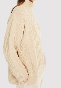 Beige neuletakki korkealla kauluksella, jossa on palmikkoneulosta, tekstuurisia yksityiskohtia ja resorit hihansuissa. Laiska istuvuus ja sivutaskut.