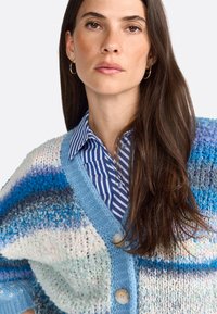 Cárdigan de punto en tonos multicolores de azul y blanco, con cuello en V, grandes botones y una textura suave, superpuesto a una camisa a rayas azul.