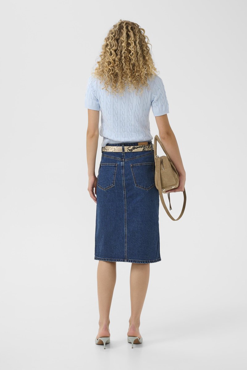 Femme aux cheveux bouclés portant un pull à manches courtes bleu clair, une jupe en jean longueur genou, des talons et tenant un sac à main beige, vue de dos.