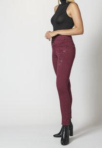 Jeans skinny burdeos con adornos, cierre de botón y bolsillos traseros. Combinados con una blusa sin mangas negra y botas de tacón negras.