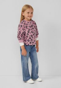 Paarse sweatshirt met zwart luipaardprint, witte manchetten, gecombineerd met lichtblauwe flared jeans en witte sneakers. Kind staat te glimlachen.
