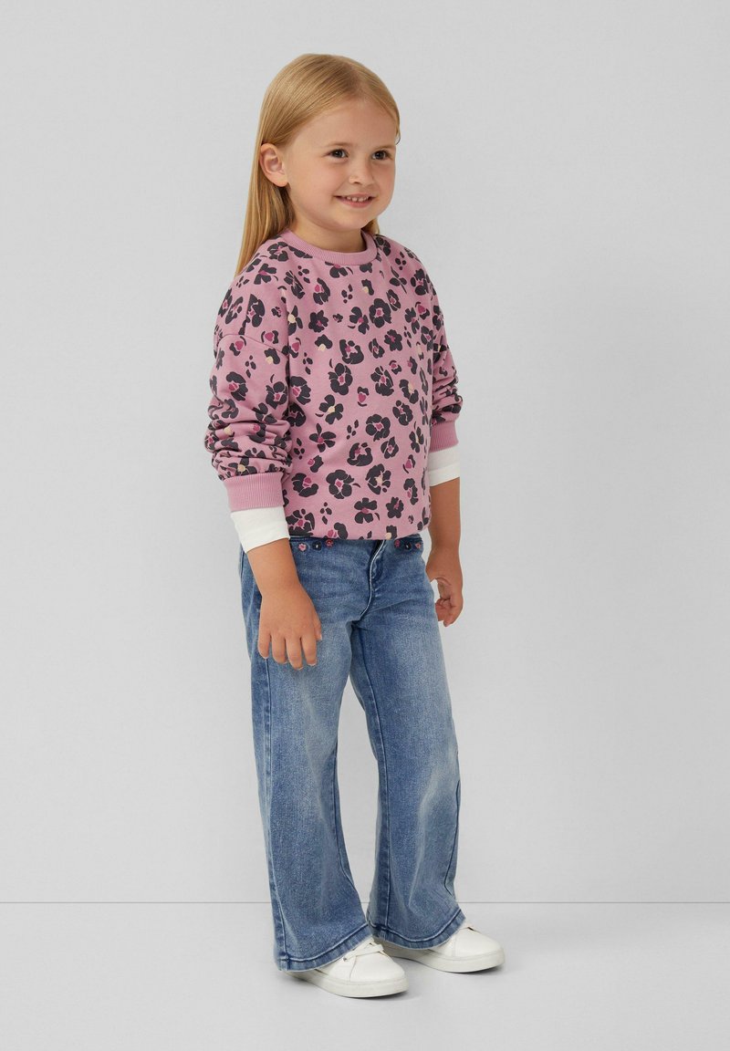 Paarse sweatshirt met zwart luipaardprint, witte manchetten, gecombineerd met lichtblauwe flared jeans en witte sneakers. Kind staat te glimlachen.