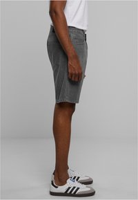 Pantalones cortos de mezclilla grises con corte recto, mostrados de perfil, combinados con zapatillas blancas y negras. Se pueden ver costuras planas y presillas para el cinturón.