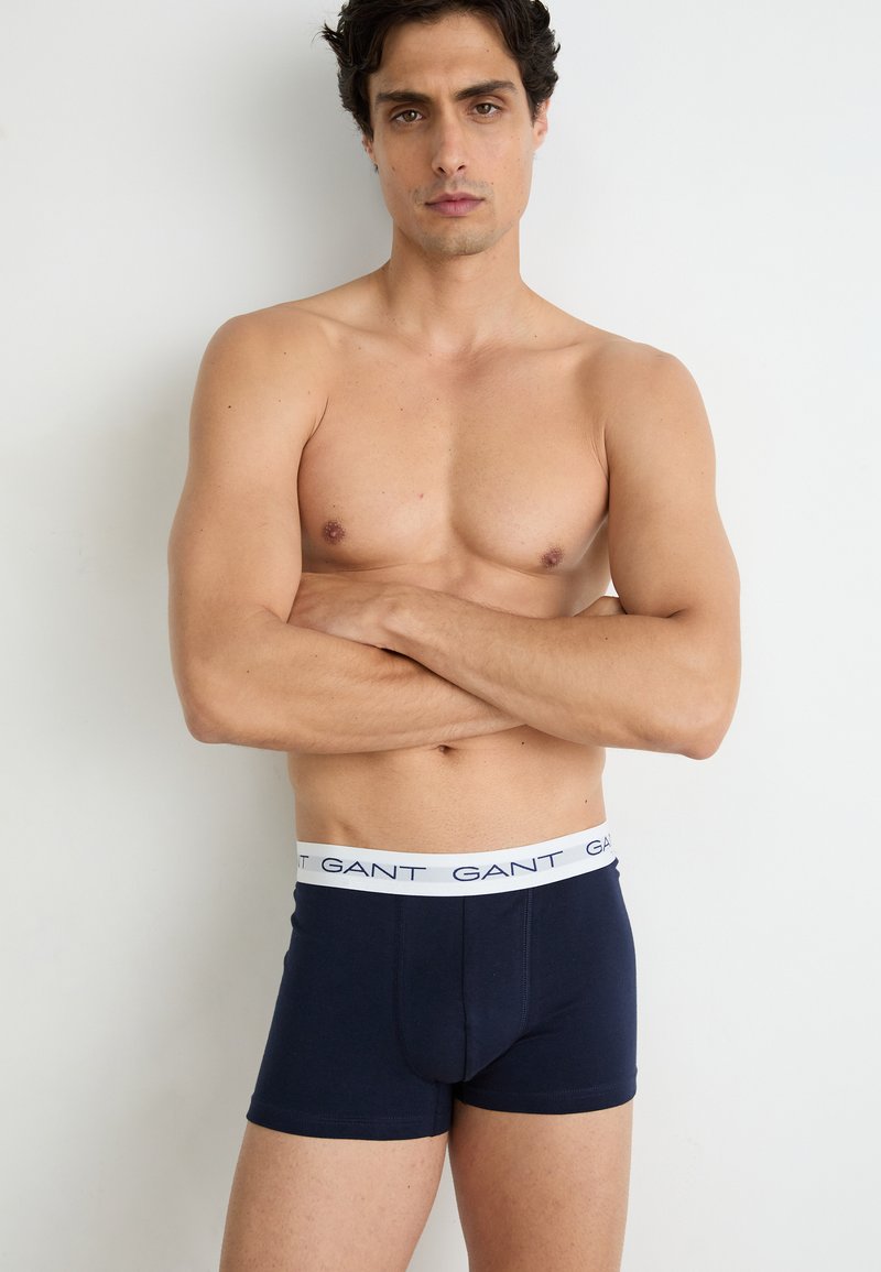 Marineblaue Boxer-Shorts mit einem weißen elastischen Bund, auf dem der Markenname steht. Modell steht mit übergeschlagenen Armen, der nackte Oberkörper ist sichtbar.