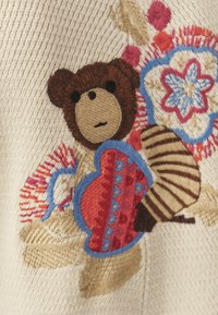 Hímzett bézs szövet, amelyen egy barna medve látható, amint egy piros szívet tart, körülötte színes virágmintákkal, az egész anyag texturált részletekkel díszítve.