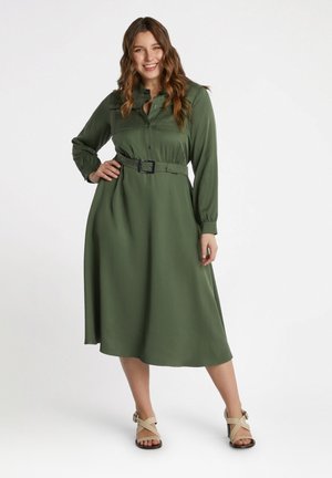 Vestito camicia verde con maniche lunghe, vita con cintura e abbottonatura frontale. Tessuto morbido e design di lunghezza midi. Abbinato a sandali di colore chiaro.