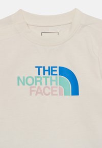 Camiseta color crema con un diseño de logo multicolor que incluye texto en azul, verde y rosa, y una forma arqueada. Material de algodón suave.