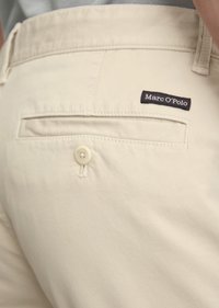 Close-up van een beige Marc O'Polo broekzak aan de achterkant met een knoop en label op een stoffen textuur.