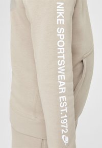 Sweatshirt bej de la Nike, cu un material texturat, mansete ribbate și un logo alb vertical pe mâneca stângă.
