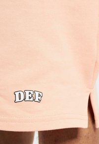 Shorts en coton de couleur pêche avec une petite fente sur le côté et un graphique noir "DEF" imprimé près de l'ourlet. Texture lisse, design décontracté.