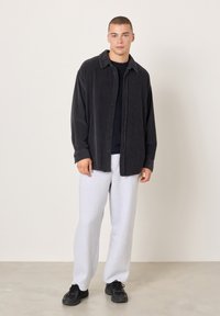 Mladić stoji, nosi crnu kordurovu košulju, crnu majicu, svijetlosive trenirke i crne tenisice, na jednostavnoj svijetloj pozadini.