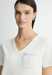 Hvid v-hals t-shirt med en struktureret lomme, der har rhinsten-detaljer. Blødt stof med en afslappet pasform, korte ærmer, minimal design.
