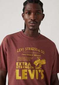 Bordeaux katoenen t-shirt met gele grafische tekst en illustratie, voorzien van een ronde hals en korte mouwen. Details bevatten de merknaam en het logo.