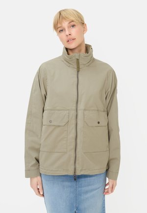 Kvinde iført beige lynlåstrøje med høj krave og forlommer med klap, kombineret med lyseblå denimnederdel, mod hvid baggrund.