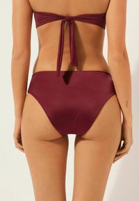 Parte inferior de bikini burdeos con diseño de pierna de corte alto, textura suave y cobertura mínima. Se ata en la parte trasera para un ajuste ajustable.