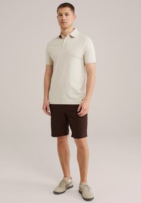 Beige polo shirt with a textured pattern and a classic collar, paired with brown shorts and light sneakers, standing on a neutral background.