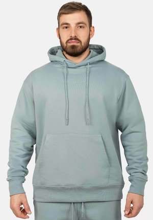 Mann mit Bart, der ein hellblaues Kapuzen-Sweatshirt mit Fronttasche und Kordeln trägt, steht vor einem schlichten Hintergrund.