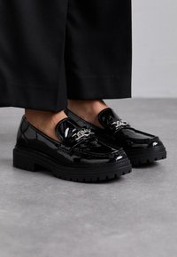 Mocassins en cuir verni noir avec une semelle épaisse en caoutchouc noir, ornés d'une chaîne décorative argentée à l'avant et d'une finition lisse et brillante.