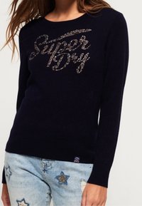 Laivastonsininen neule pyöreällä pääntiellä, jossa teksturoitu hopeinen "Super Dry" -logo etuosassa. Yhdistetty vaaleaan farkkuun, joka on koristeltu tähdillä.