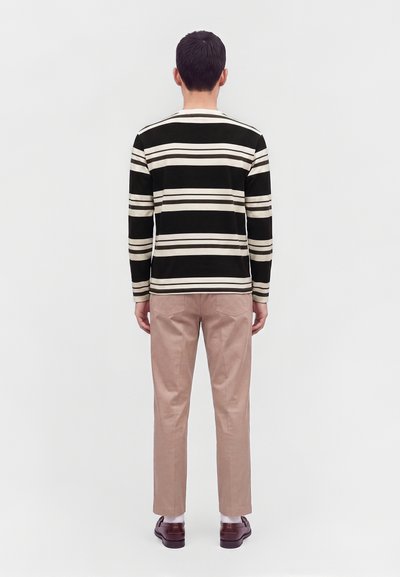Homme debout de dos portant un t-shirt à manches longues rayé noir et blanc, un pantalon beige, des chaussettes blanches et des chaussures marron.