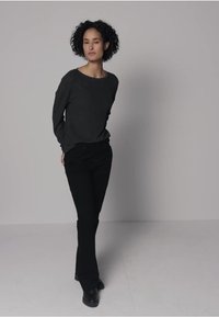 Langärmliger anthrazitfarbener Pullover, kombiniert mit schwarzen Schlaghosen und schwarzen Schuhen, getragen von einem Modell vor einem hellen Hintergrund.