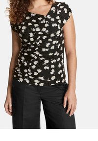 Top corto negro de manga corta con patrón floral blanco, que presenta un detalle fruncido en la parte delantera y un escote en forma de V. Se lleva con pantalones negros.