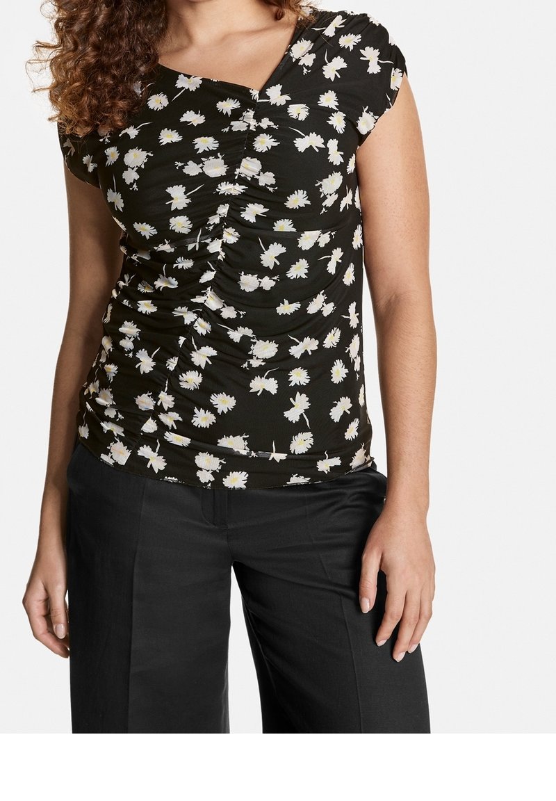 Top corto negro de manga corta con patrón floral blanco, que presenta un detalle fruncido en la parte delantera y un escote en forma de V. Se lleva con pantalones negros.