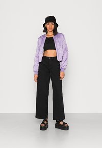 Casaco bomber de cetim roxo com um design curto, combinado com calças largas pretas de cintura alta e sandálias pretas de plataforma. Acessório de chapéu balde preto.