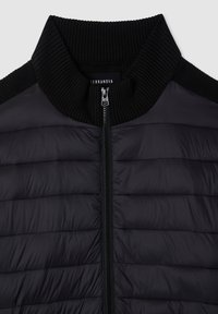 Giacca a vento nera con colletto a costine, chiusura frontale a zip completa e texture trapuntata. Il materiale appare lucido con motivi di cucitura orizzontali.