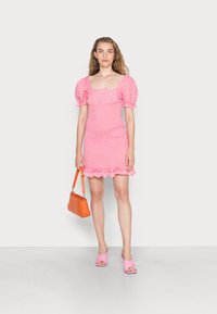 Vestido rosa de decote quadrado com mangas bufantes e bainha com pregas, segurando uma bolsa laranja. Combinado com sapatos de salto com tiras cruzadas rosa. Textura suave.