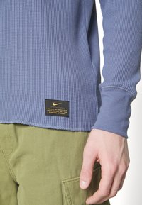 Gros plan sur une personne portant une chemise à manches longues bleue texturée avec une étiquette Nike noire et un pantalon vert olive avec une poche visible.