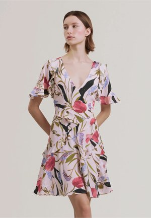 Donna che indossa un vestito floreale rosa chiaro con scollo a V profondo e maniche corte arricciate, in piedi con le mani dietro la schiena su uno sfondo semplice.