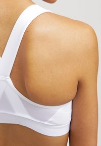 Vista posteriore di una persona che indossa un reggiseno sportivo bianco con pannelli in rete, che mostra la pelle nuda della spalla e della parte superiore della schiena.