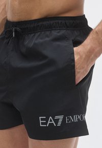 Fekete fürdőnadrág könnyű szövetből, gumis derékrésszel és húzózsinórral, oldalzsebekkel és ezüst EA7 Emporio Armani logóval.