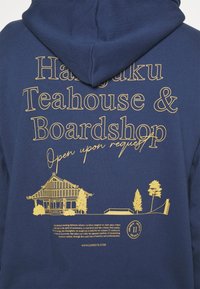 Felpa navy con testo giallo "Harajuku Teahouse & Boardshop Apertura su richiesta," una pagoda, alberi, un ramp e un logo rotondo.