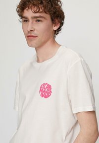 Weißes Baumwoll-T-Shirt mit einem kreisförmigen pinken "ESCAPEMOOD"-Grafikbereich auf der linken Brust, kurzen Ärmeln und Rundhalsausschnitt.