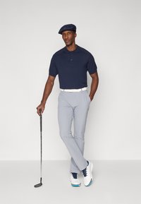 Marine polo-shirt, lyse grå golfbukser, hvit belte og hvite sko med marine detaljer, stående med en golfklubbe. Flat caps på hodet.
